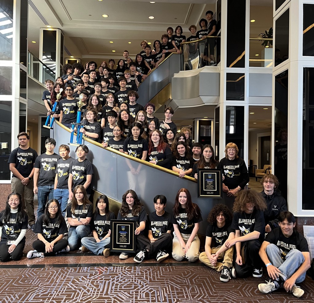 Group photo of all musicians and Director Tyra Cable wearing black AHS Music tee shirts in a lobby, holding trophies and plaques