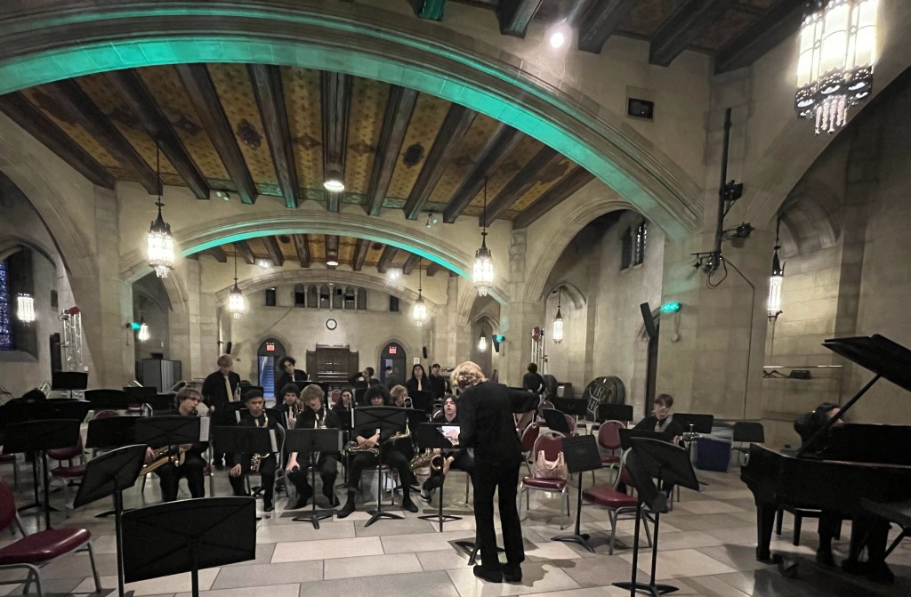 A jazz band rehearses in an old church hall with wooden beans and stone arches. The musicians and music director are wearing all black, and musicians also have straight gold ties
